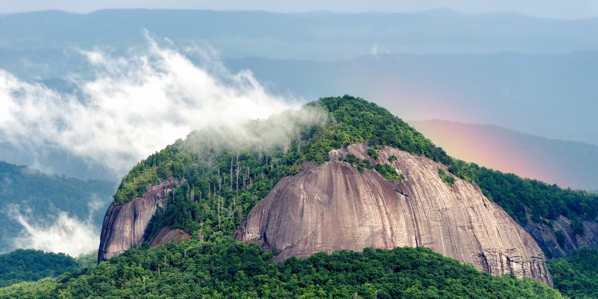 Large rock outcroping shrouded in clouds with a faint rainbow and mountains behind. Taken from the Blue Ridge Parkway.